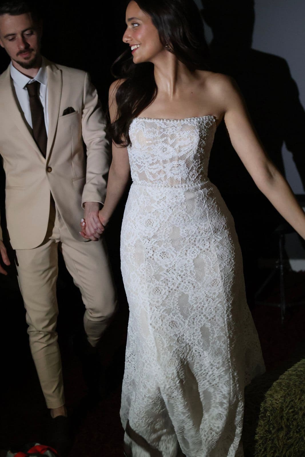 A bride in a lace gown walking hand in hand with her partner at night
